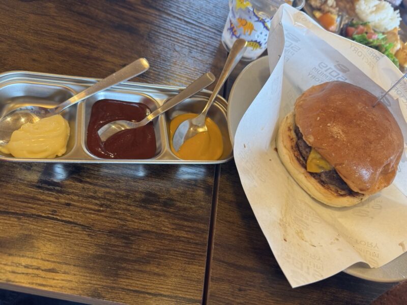 チーズバーガーが紙に包まれて皿に盛られ、横には3種類のソース（マスタード、ケチャップ、マヨネーズ）が入った小分けトレイが置かれている