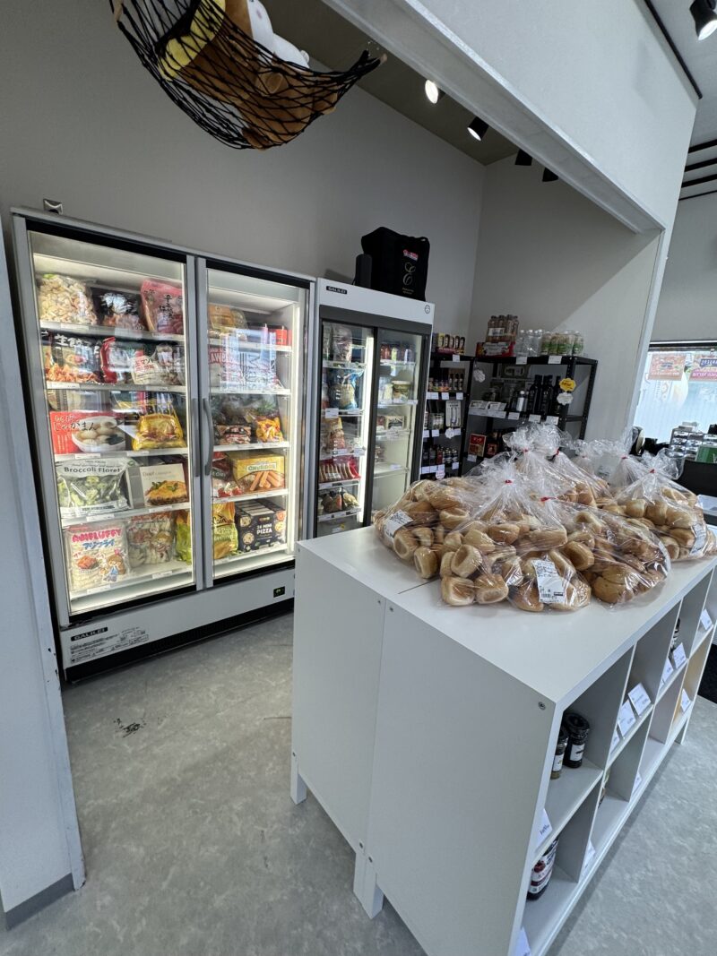 メゾンクロス店内の冷凍食品コーナーとベーカリー商品。左側には冷凍ショーケースが3台設置され、冷凍ピザやブロッコリー、チキンナゲットなどの冷凍食品が並んでいる。右側の白いカウンターには透明な袋に包装されたディナーロールやベーグルなどのパン類が陳列されている。天井には黒いネットハンモックが吊るされている。