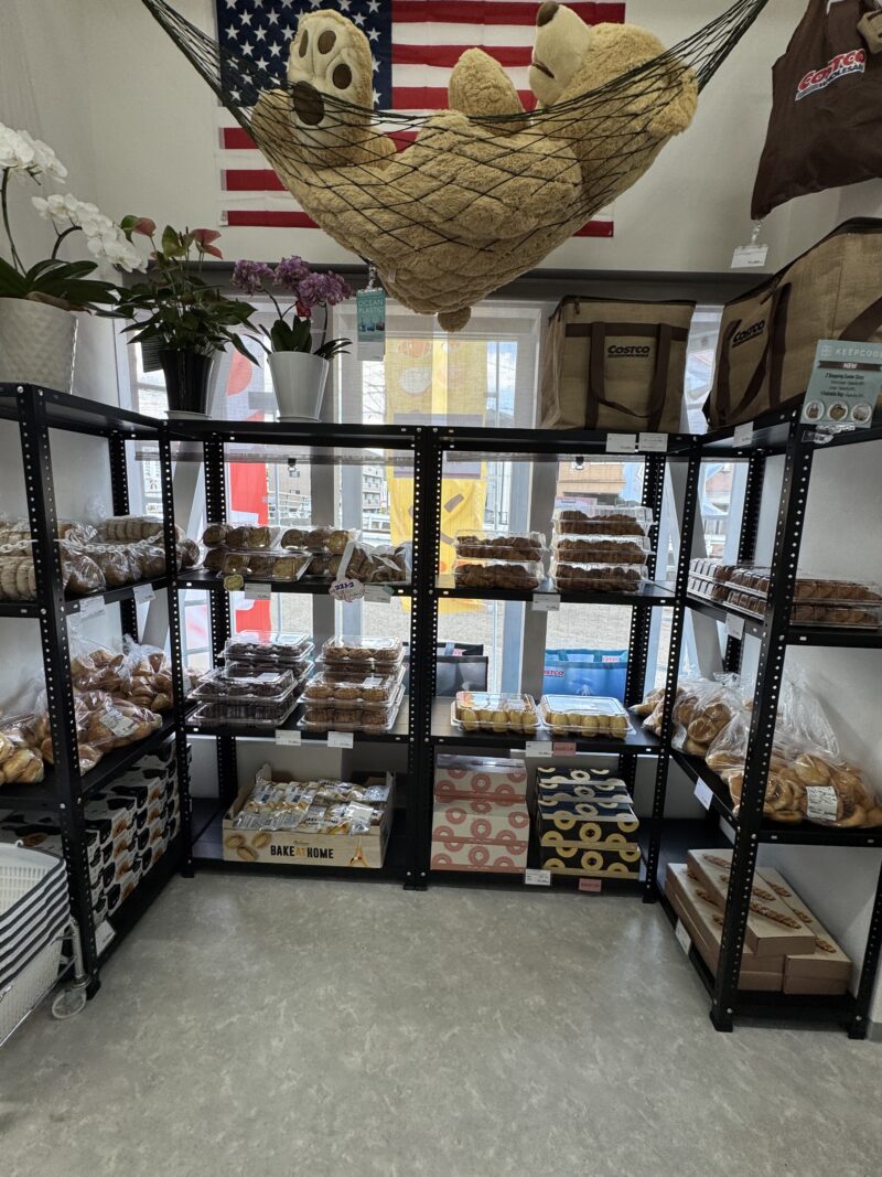 メゾンクロス店内のベーカリーコーナー。黒いスチールラックに透明パッケージに入ったパン類やスイーツが並んでいる。上部には大きなテディベアがハンモックに入れられ、アメリカ国旗をバックに飾られている。窓の側にはコストコのエコバッグと観葉植物が置かれている。