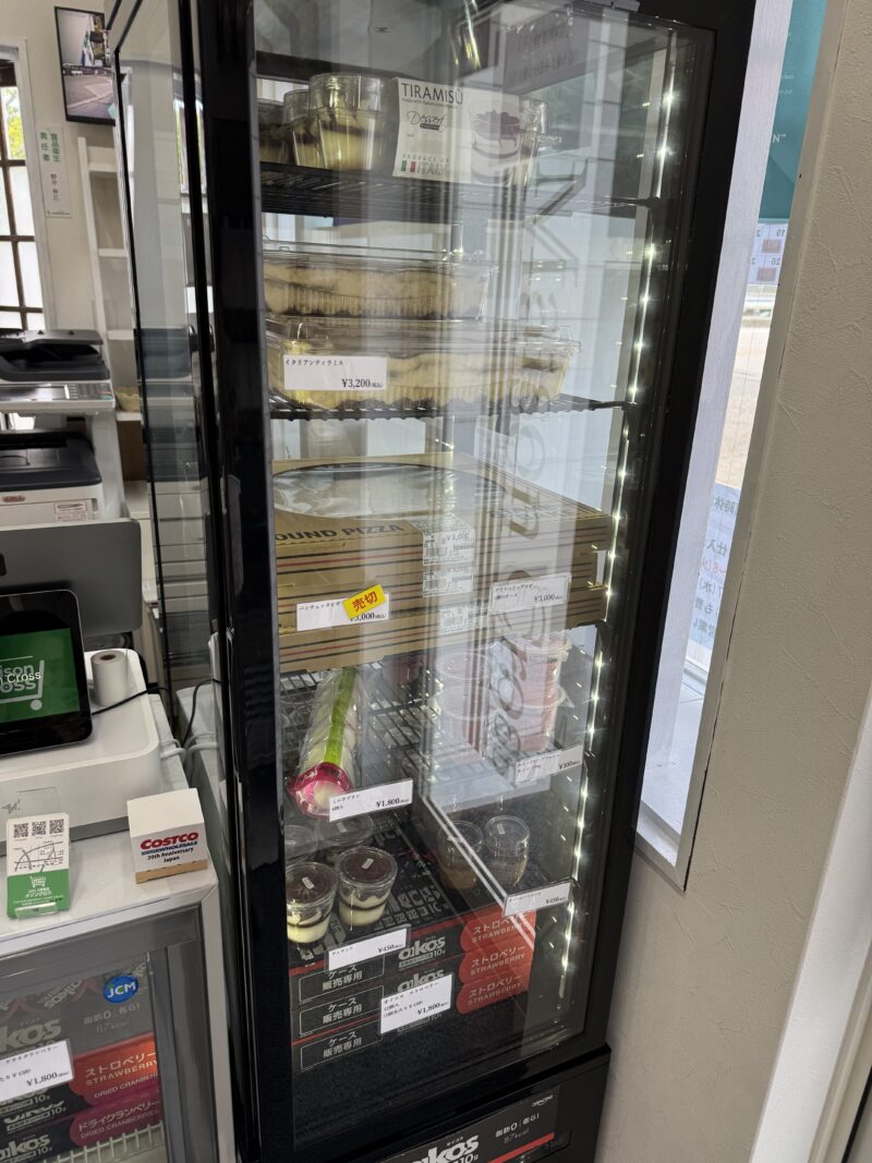 メゾンクロス店内の冷蔵ショーケース。ガラス扉を通して、ティラミスなどのデザート、ピザ、オイコスヨーグルトなどが陳列されている。価格表示も見え、上段にはティラミス(3,200円)、中段には「売切」表示のあるピザ(3,000円)、下段にはヨーグルトなどの商品が並んでいる。