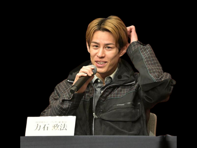 力石政法選手が記者会見で、マイクを持ちながら頭に手をやる仕草をしている様子。黒い背景に、チェック柄のシャツの上に黒いジャケットを着用し、前には「力石政法」と書かれた名札が置かれている。
