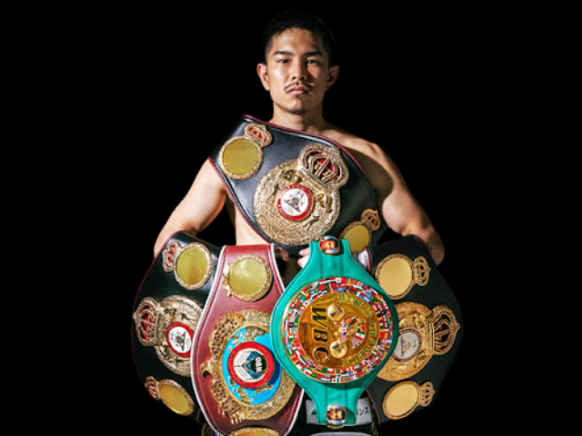 黒い背景の前で立つ井岡一翔選手の公式ポートレート写真。上半身裸で、WBA、WBC、WBOなど複数の団体の世界チャンピオンベルトを胸の前に持ち、複数の階級での世界王者としての実績を示している。