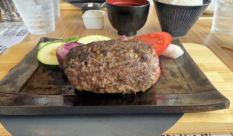 熱々の鉄板で提供される「たちばなのバーグステーキ」の塩バーグステーキ。分厚いハンバーグが野菜と一緒に盛り付けられ、背景にはご飯と味噌汁が見える。