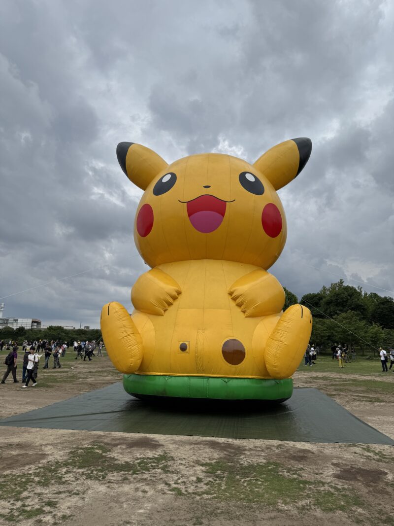 万博記念公園に設置された巨大なピカチュウのエアバルーン。黄色い体に赤いほっぺ、黒い耳先が特徴的で、緑のマットの上に座っている。曇り空の下、多くの来場者が周囲で写真撮影をしている。