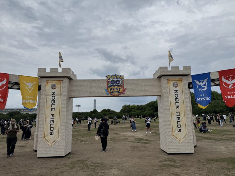 Pokémon GO Fest 2025大阪のメインエントランスゲート。石造り風の城門デザインで、中央にGO Festロゴ、両脇に「NOBLE FIELDS」の文字とPokémon GOの3チーム(インスティンクト、ミスティック、ヴァーラー)のバナーが掲げられている。太陽の塔が背景に見え、多くの参加者が行き交っている。