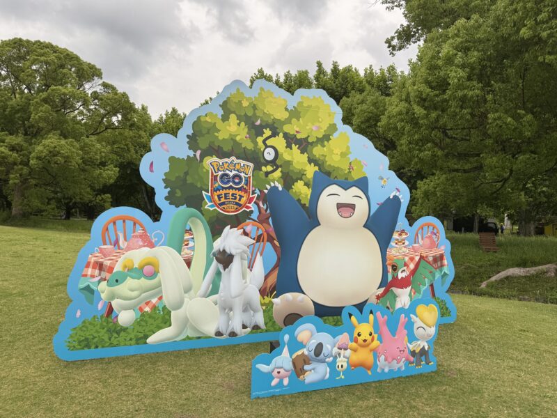 Pokémon GO Fest 2025大阪のピクニック風フォトスポット。大きな木の下でカビゴンが手を広げて座り、周囲にライチュウ、レシラム、ポッチャマ、ピカチュウなど様々なポケモンが配置された立体的なパネル装飾。前面には小さなポケモンたちのパネルも設置されている。