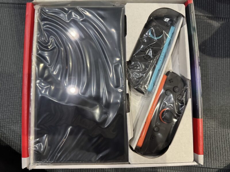 Nintendo Switch 2の開封画像。箱の中に黒いビニール袋に包まれた本体と、右側にJoy-Con 2(青とオレンジ色)が個別包装されて配置されている