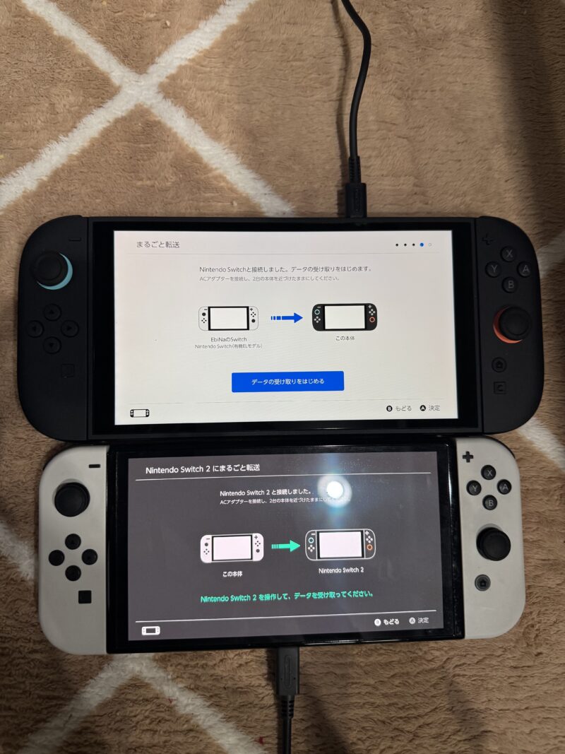 Nintendo SwitchからSwitch 2へのデータ移行画面が表示された2台の本体。上がSwitch 2で「まるごと転送」画面、下がSwitch有機ELで「Nintendo Switch 2にまるごと転送」画面が映っている