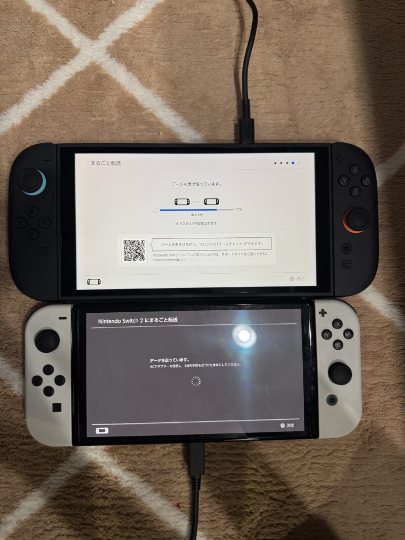 Nintendo Switch 2のデータ移行進行中の画面。上のSwitch 2には77%完了を示すプログレスバーとQRコード、下のSwitch有機ELには「データを送っています」というメッセージとローディングアニメーションが表示されている